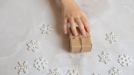 a small childs hand reaches for a Christmas gift packed in crab packaging on a white wooden background with snowflakes of their treeの写真素材