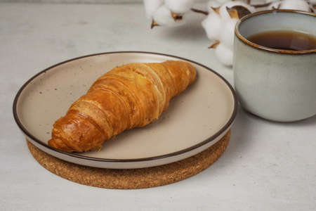 croissant on a beige plate with a cup of tea and a sprig of cotton on a concrete backgroundの写真素材
