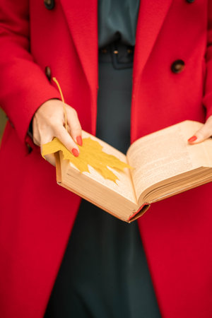 A European girl in a red coat holds an open book with a yellow maple leaf in her handsの写真素材