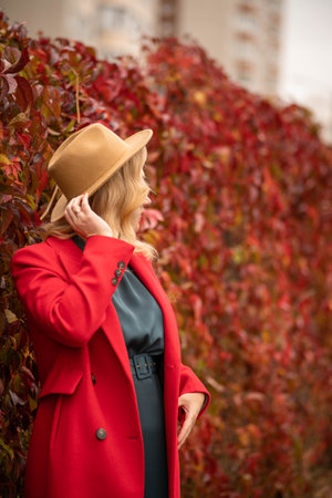 A European girl in a red coat with a beige hat on a background of red leaves looks away, the concept of golden autumnの写真素材