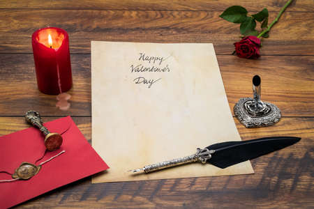 Lovely classic Valentineâs Day cad, hand written with ink and quill on vintage paper, red envelope with wax seal, red rose, lit candle, silver quill stand on antique oak â front viewの写真素材