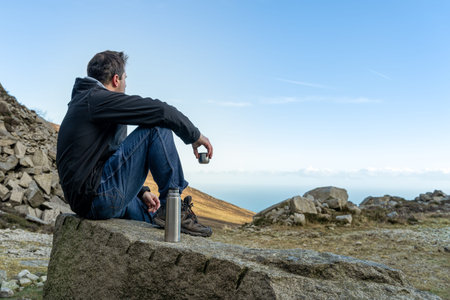 Middle age man sitting on the rock drinking a tea or coffee in cold morning looking on the vale and sea from mountain range, view from behindの写真素材