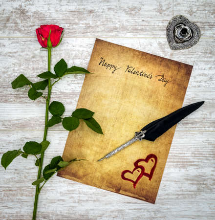 Antique preachment with hand written Happy Valentineâs Day, red rose, red painted 2 harts, detailed silver quill stand in shape of hart accented by flat lying beautifully ornamented silver and black quill on rustic white painted oakの写真素材