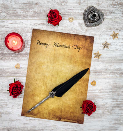 Antique preachment with hand written Happy Valentineâs Day, red lit candle, 3 red roses, wooden decorations,  detailed silver quill stand in shape of hart accented by flat lying beautifully ornamented silver and black quill on rustic white painted oakの写真素材