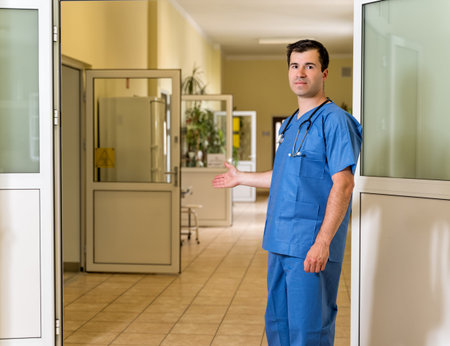 Middle age, Caucasian white, male doctor in blue scrubs with stethoscope inviting patients in to the clinic or hospital, blurred hospital corridor in backgroundの写真素材