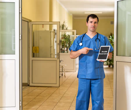 Middle age, Caucasian white, male doctor in blue scrubs with stethoscope holding and pointing to tablet with Health Insurance text, blurred hospital corridor in backgroundの写真素材
