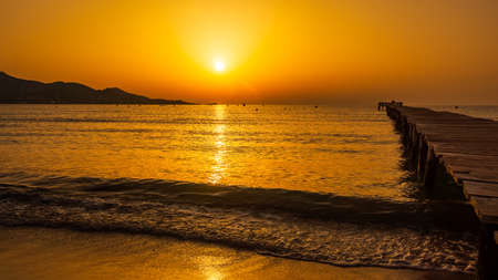 Orange solar disc in sunrise on beach with silhouette of mountains and pier on bay, reach orange and yellow colours of the sky panorama Mallorca Spainの写真素材