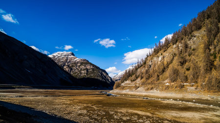 Beautiful mountain valley with stream and riverbed, trees and dried Livigno lake, puffy clouds and...