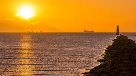 Silhouette of cargo ships at golden hour. Sun, solar disc, shines from behind cloud. Lighthouse and breakwater on Bull Island, Dublin, Irelandの写真素材