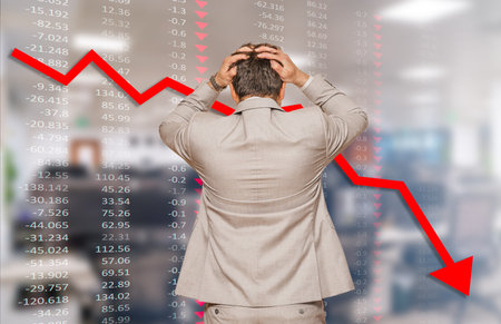 Back view of middle age businessman pulling his hair out as his business falling during recession, data chart and blurred office in backgroundの写真素材