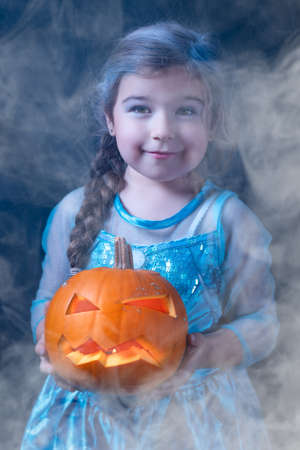 Halloween portrait of young girl stylised for frozen princess in a blue dress. Smiling child holding orange curved pumpkin surrounded by smoke or mistの写真素材
