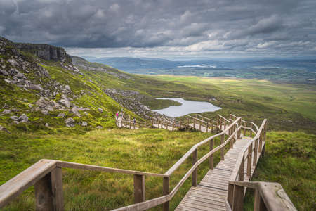 People enjoying a walk on steep stairs of wooden boardwalk in Cuilcagh Mountain Park with a view on lake and valley below, Northern Irelandの写真素材