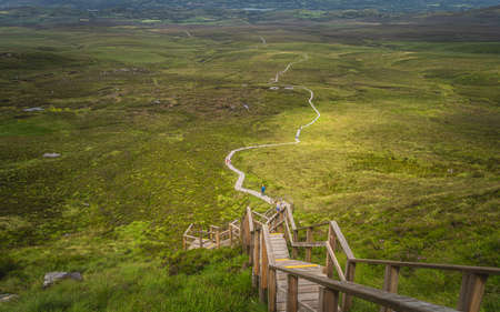 People climbing on steep stairs of wooden boardwalk, to reach Cuilcagh Mountain peak with view on green fields and valley below, Northern Irelandの写真素材