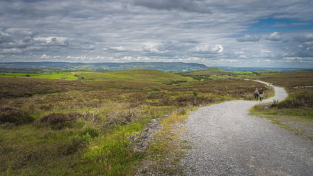 People walking on the trail between green hills and fields with stormy, dramatic sky in background,...