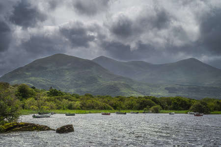 Wooden boats moored on Lough Leane with mountains of Killarney National Park and dark dramatic clouds in background, Ring of Kerry, Irelandの写真素材