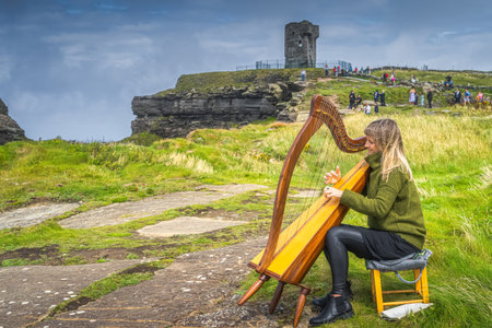Doolin, Ireland, August 2019 Woman playing harp on the top of iconic Cliffs of Moher, popular...