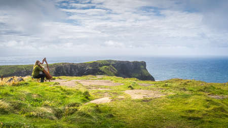 Woman playing harp on the top of the cliff with stunning view on iconic Cliffs of Moher, popular tourist attraction, Wild Atlantic Way, Clare, Irelandの写真素材