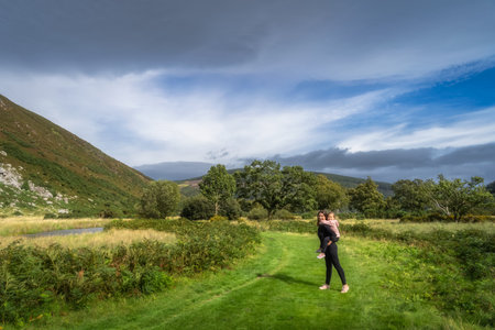 Young girl piggyback riding on her mum on a country road with river and green forest in background. Family hike, Lough Dan, Wicklow Mountains, Irelandの写真素材