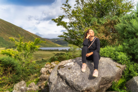Young woman sitting on a large rock and smiling. Lake Lough Dan and mountain valley in background,...