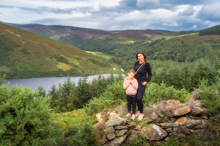 Mother and daughter standing and smiling on rock wall. Lake Lough Dan, forest and fields in...