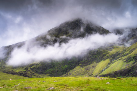 A breathtaking view of a majestic Carrauntoohil mountain elegantly shrouded in fluffy clouds, surrounded by vibrant green hills that stretch under a dramatic, colorful sky, Kerry, Irelandの写真素材