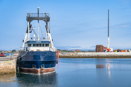 Moored blue fishing trawler at harbor in Galway, Rossaveel, Ireland. Hull reflection on calm water. Quay and breakwater in background, dockside crane and containers.の写真素材