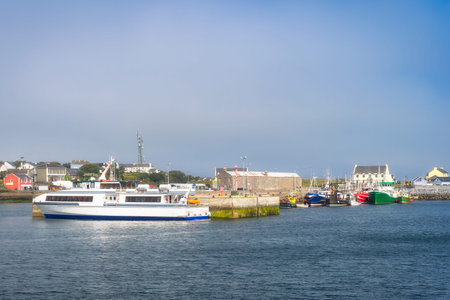 Coastal ferry nearing Inis Mor harbor with village skyline in background, Galway in Ireland. Mixed fleet at quay, commuter and leisure transport vibe, soft light over rippling waterの写真素材