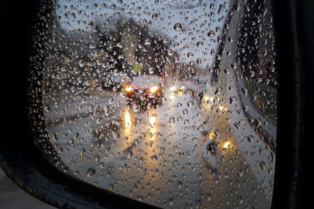Reflection of cars with headlights on in the wing mirror of a car with raindrops. Reflection of cars with headlights on in the wing mirror of a car with raindrops. Car traffic on a city street in the rain.の写真素材