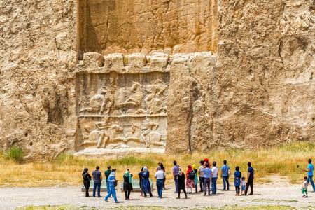 NAQSH-E RUSTAM, IRAN - MAY 3, 2015: The group of tourists with the tour guide checking out the relief of the ancient necropolis near ruins of old city Persepolis.のeditorial素材