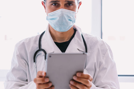 Black haired doctor in white coat and stethoscope looking at his tablet, in a window. Medicine...