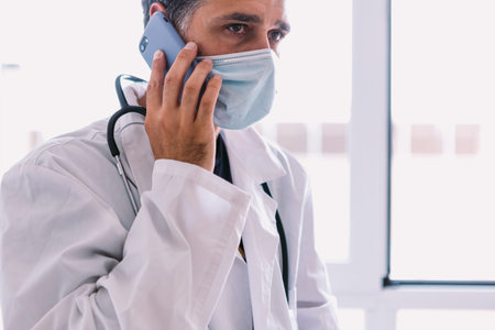 Black haired doctor in white coat and stethoscope talking on his mobile phone, at a window. Medicine...