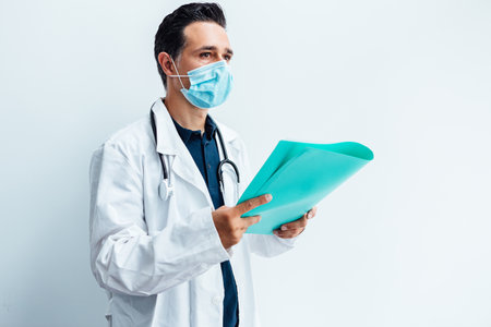 Black haired doctor wearing white coat and stethoscope looking at his mobile phone and smiling while making a video call, on white background. Medicine conceptの写真素材