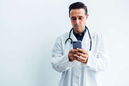 Black haired doctor wearing white coat and stethoscope looking at his mobile phone and smiling while making a video call, on white background. Medicine conceptの写真素材