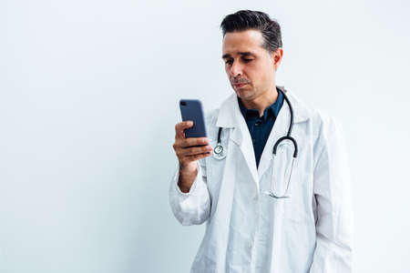 Black haired doctor wearing white coat and stethoscope looking at his mobile phone and smiling while making a video call, on white background. Medicine conceptの写真素材