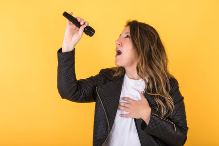 Young singer girl, wearing black leather jacket and microphone, making surprise gesture on yellow...