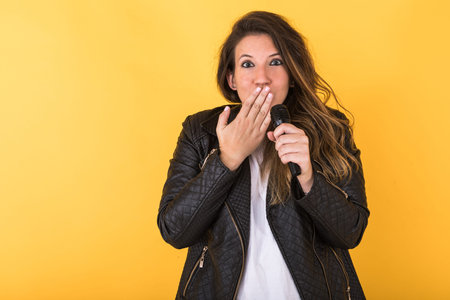 Young singer girl, wearing black leather jacket and microphone, making surprise gesture on yellow...