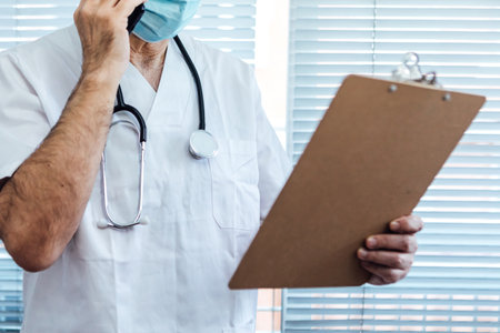 Mature male doctor - nurse wearing face mask, talking on mobile phone and holding a report next to a hospital window medicine conceptの写真素材
