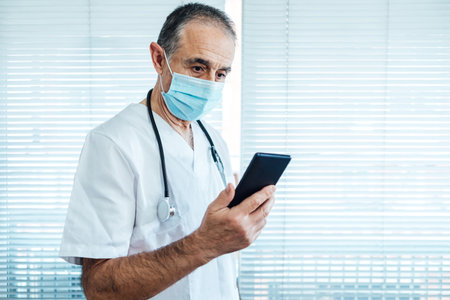 Mature male doctor - nurse wearing face mask, talking on mobile phone and holding a report next to a hospital window medicine conceptの写真素材