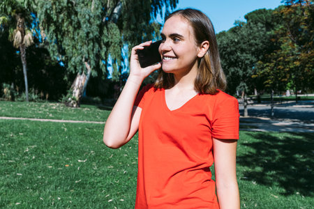 Young blonde wearing a red shirt looks at the cell phone and listens to music in a park, leaning on...
