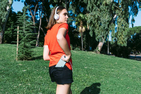 Young blonde wearing a red shirt looks at the cell phone and listens to music in a park, leaning on the trunk of a treeの写真素材