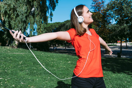 Young blonde wearing a red shirt looks at the cell phone and listens to music in a park, leaning on...