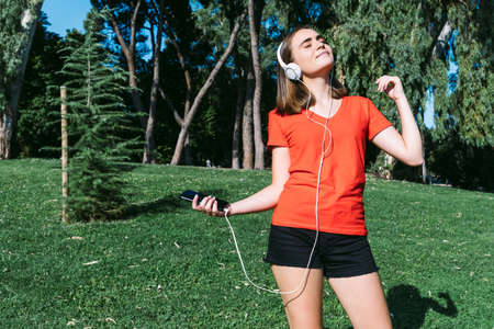 Young blonde wearing a red shirt looks at the cell phone and listens to music in a park, leaning on the trunk of a treeの写真素材