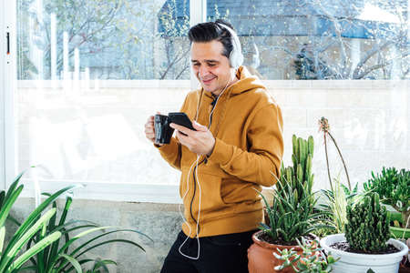 Smiling man looking at the mobile and listening to music with headphones near the window, surrounded by green plants and flowersの写真素材