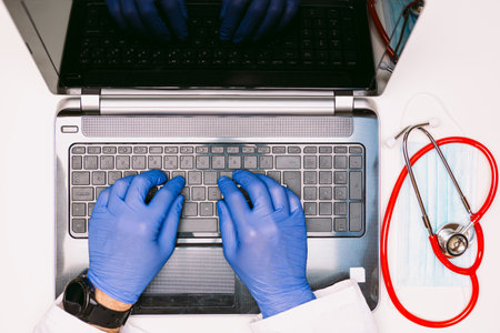 Overhead view of a laptop and the hands of a doctor with latex gloves typing on the keyboard, with a stethoscope at the aldoの写真素材
