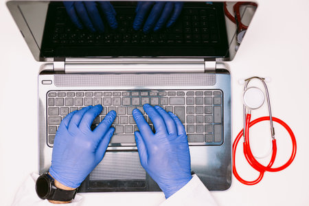 Overhead view of a laptop and the hands of a doctor with latex gloves typing on the keyboard, with a stethoscope at the aldoの写真素材