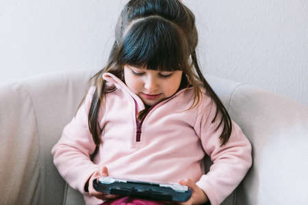 Little black haired girl wearing a pink sweater playing a video game with a portable video console on a sofaの写真素材