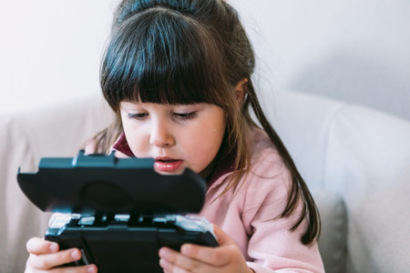 Little black haired girl wearing a pink sweater playing a video game with a portable video console...