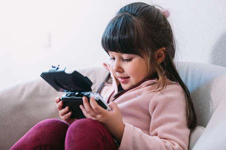 Little black haired girl wearing a pink sweater playing a video game with a portable video console on a sofaの写真素材