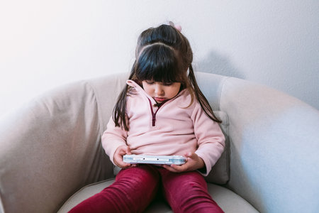 Little black haired girl wearing a pink sweater playing a video game with a portable video console on a sofaの写真素材