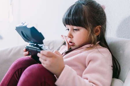 Little black haired girl wearing a pink sweater playing a video game with a portable video console on a sofaの写真素材